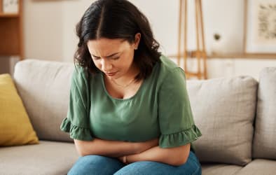 An image of a woman sitting on her couch, holding her stomach with a pained expression.
